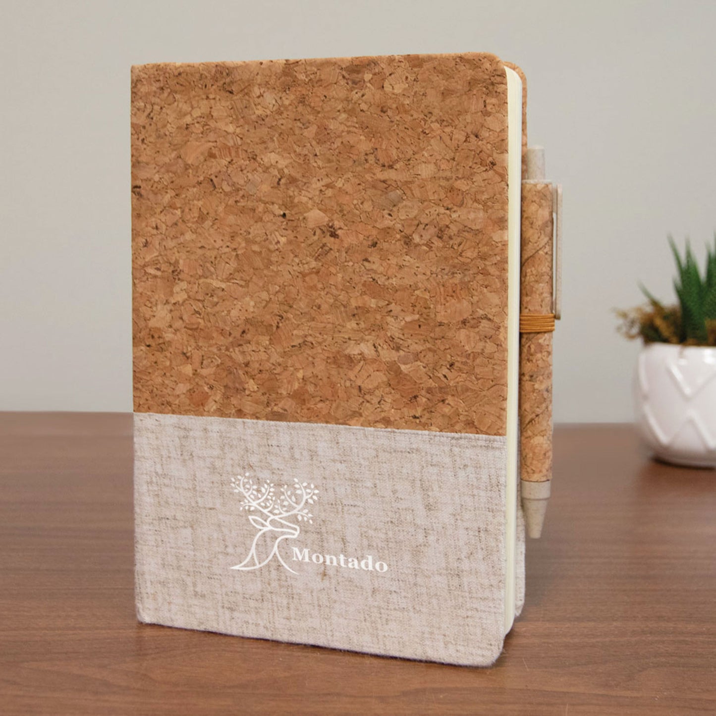 Eco-friendly Montado journal with cork top and beige linen fabric bottom, featuring a white Montado logo with a stylized tree design, and a matching cork-covered pen attached on the side.