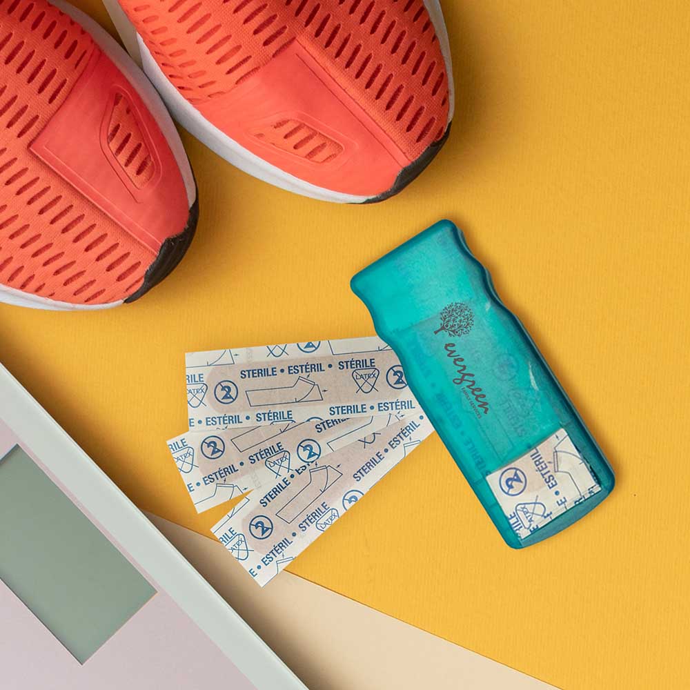 Compact teal bandage dispenser with printed logo 'evergreen' next to scattered sterile bandages, placed beside orange sneakers on a yellow surface