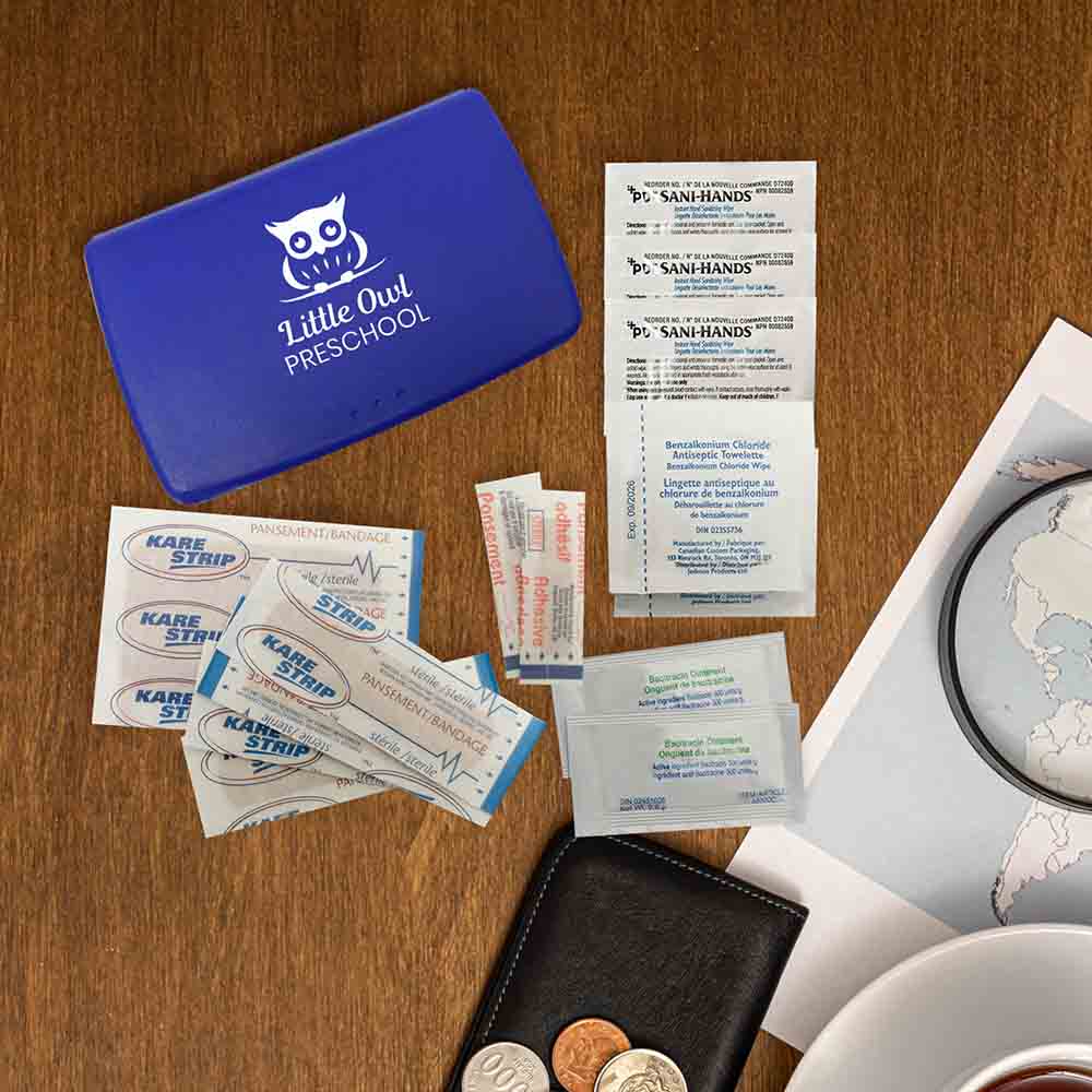 Blue first aid case with “Little Owl Preschool” logo, displayed on a wooden surface alongside bandages, ointments, and antiseptic wipes.
