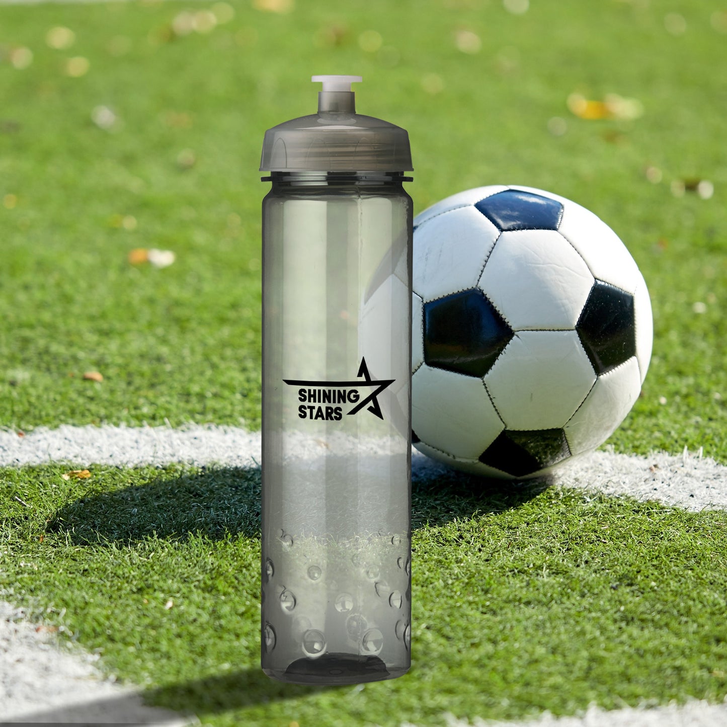 Translucent gray sports bottle with 'Shining Stars' logo placed on a soccer field next to a black and white soccer ball.