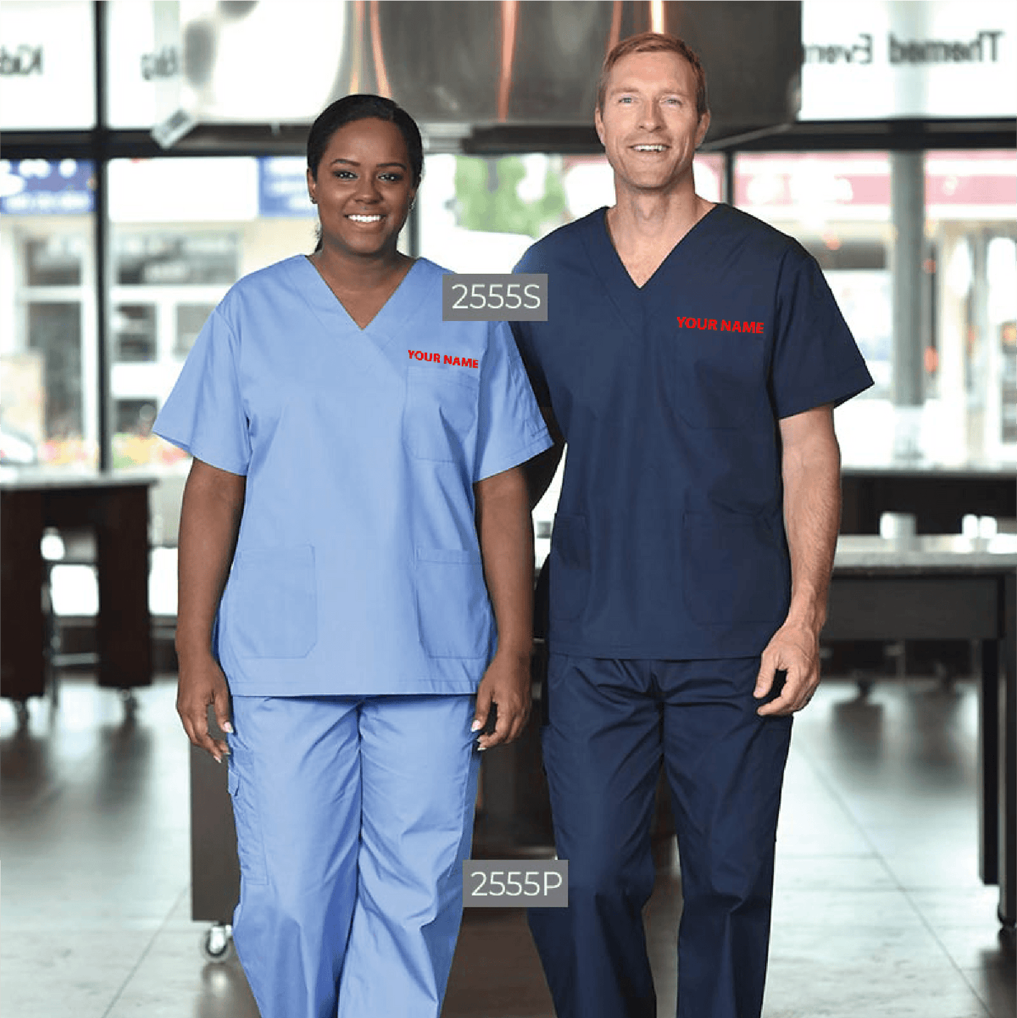 Two models wearing the Premium Unisex V-Neck Scrub Set - Logo Embroidered; the woman is in a light blue V-neck top (style 2555S) and matching pants (style 2555P), while the man wears a navy V-neck top (style 2555S) with coordinating pants (style 2555P), both featuring red embroidered 'YOUR NAME' text on the chest.