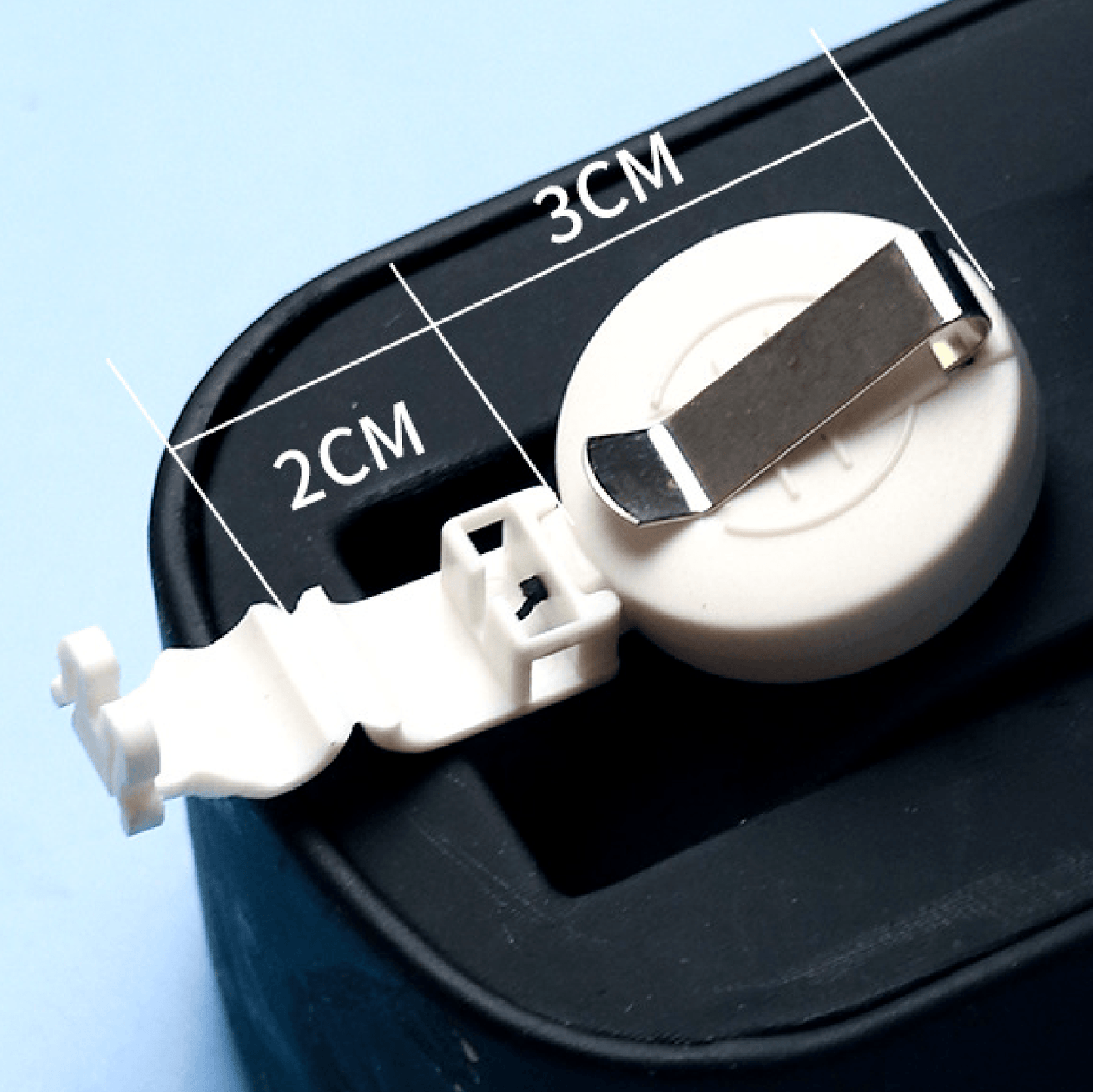 Close-up of a white retractable badge reel with metal clip, attached to a plastic mount with size measurements labeled as 2CM and 3CM, placed on a black surface with a light blue background.