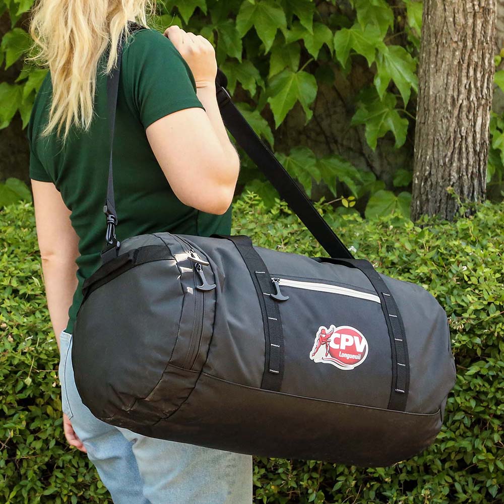 Black duffel bag worn with shoulder strap, featuring front zip pocket and outdoor setting
