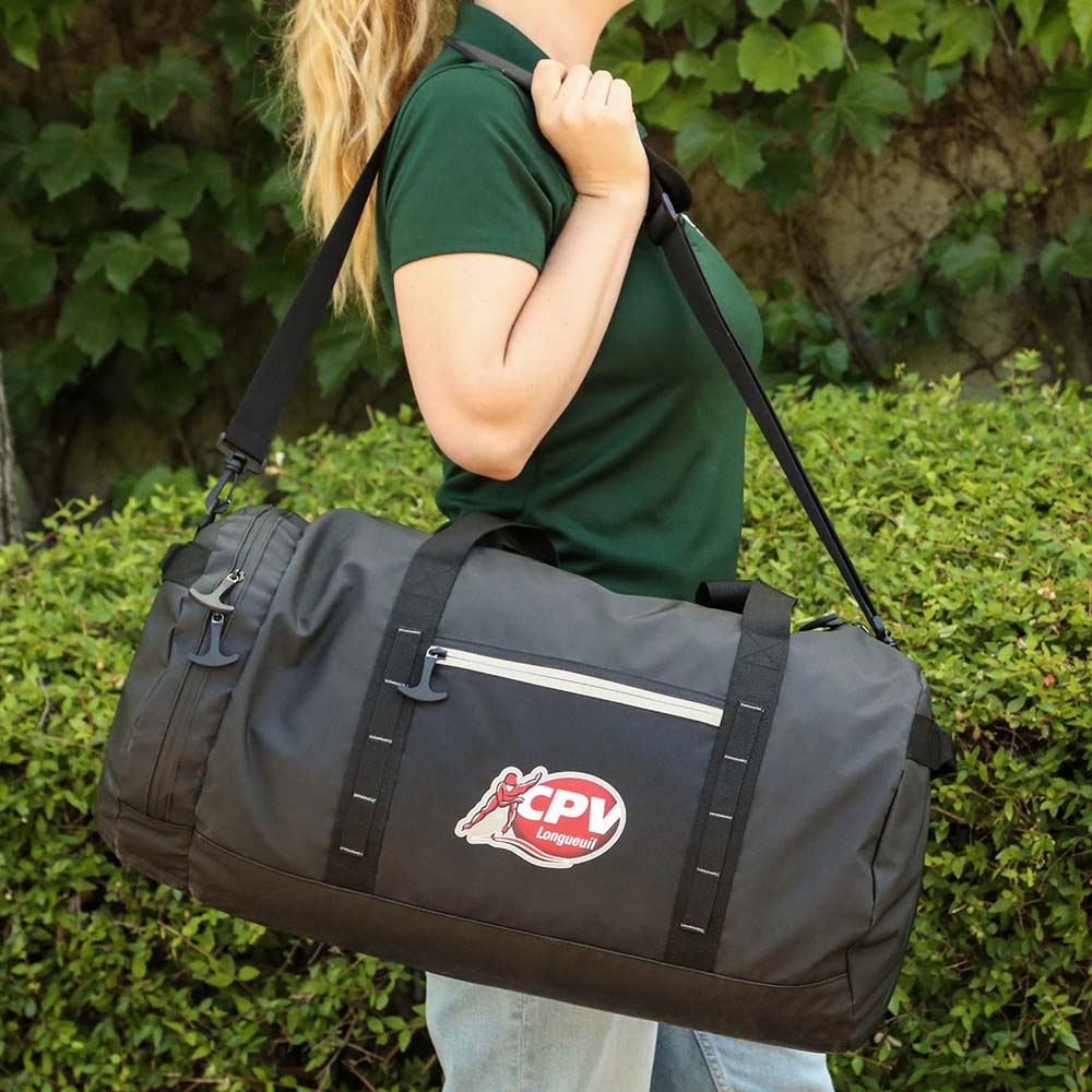 Black duffel bag worn with shoulder strap, featuring front zip pocket and outdoor setting