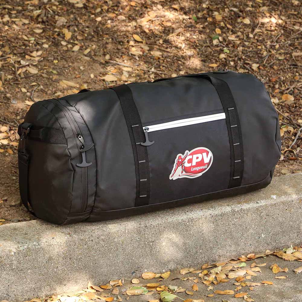 Black duffel bag worn with shoulder strap, featuring front zip pocket and outdoor setting