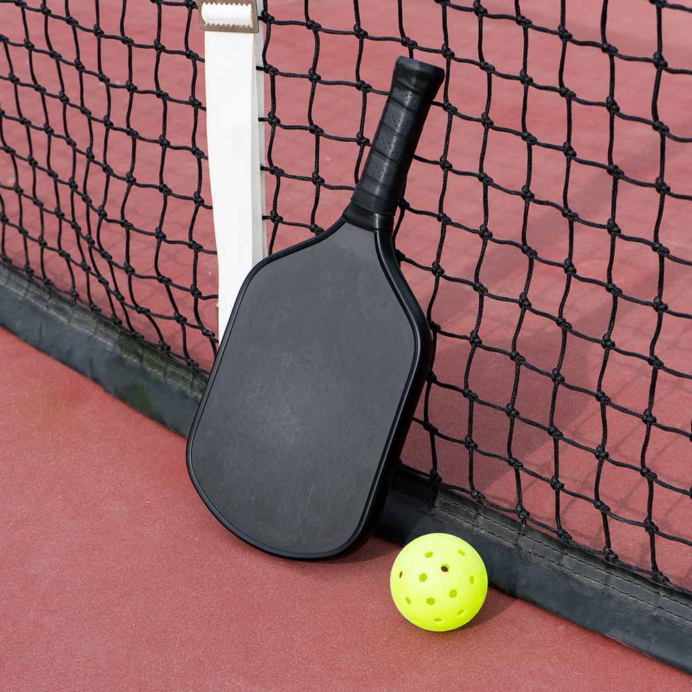 Black pickleball paddle leaning against a net on a red court surface, with a yellow perforated pickleball resting nearby.