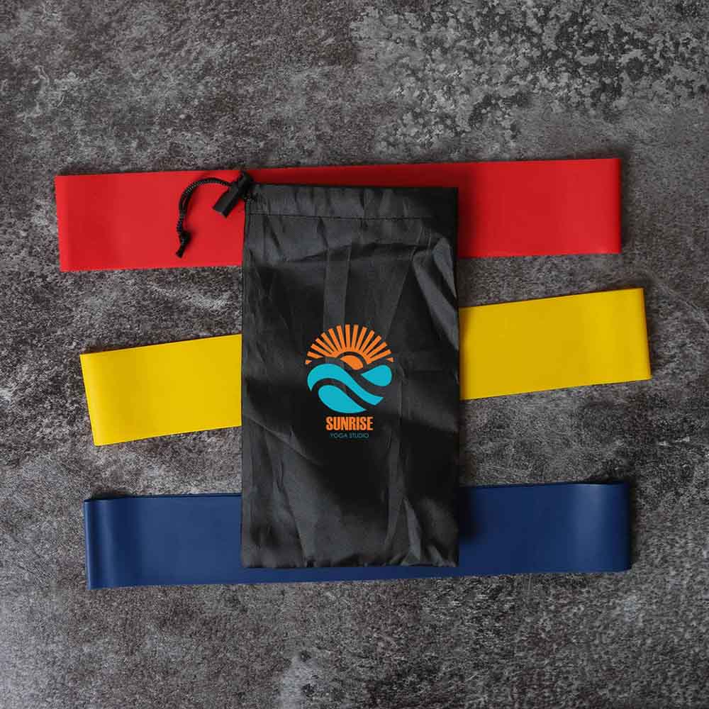 Red, yellow, and blue resistance bands laid out with a black Sunrise Yoga Studio drawstring pouch on top, set against a textured dark background.
