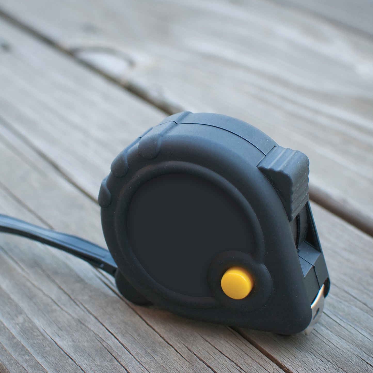 Compact black tape measure with a yellow button, placed on a wooden surface, featuring a blank circular branding area.