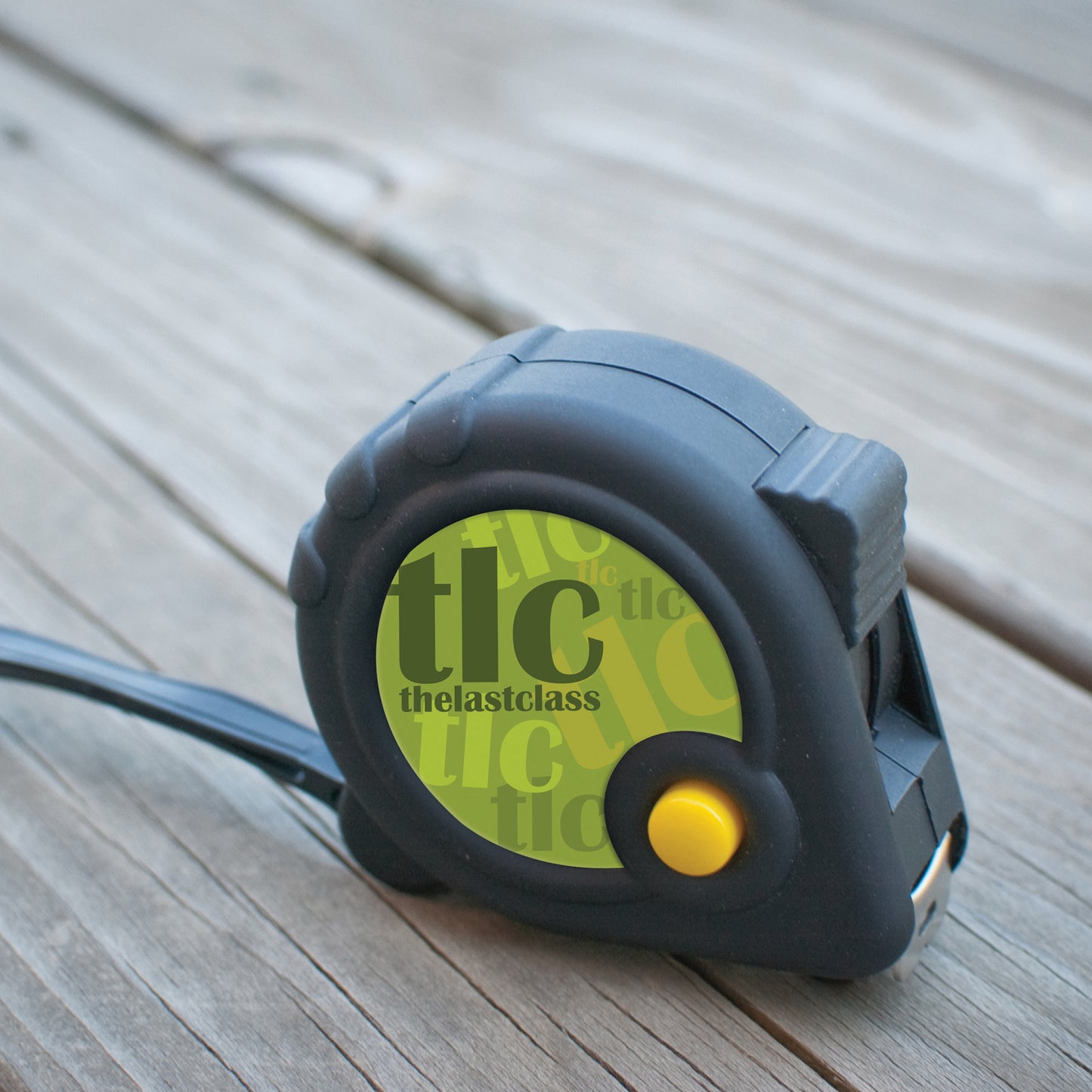Black and yellow tape measure with a green circular branding area featuring the logo, placed on a wooden surface.