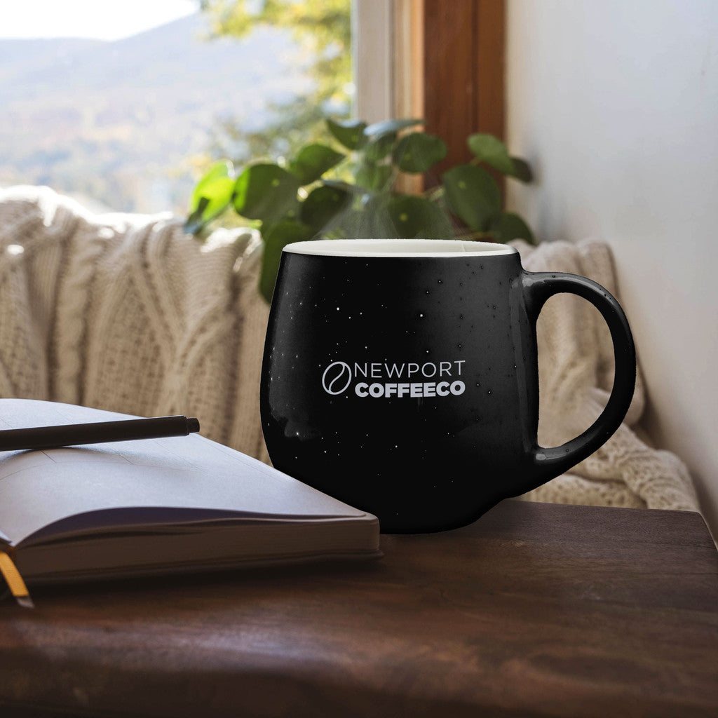 Black speckled ceramic mug with NEWPORT COFFEE CO logo, placed on a table near a window with plants and a blanket in the background.