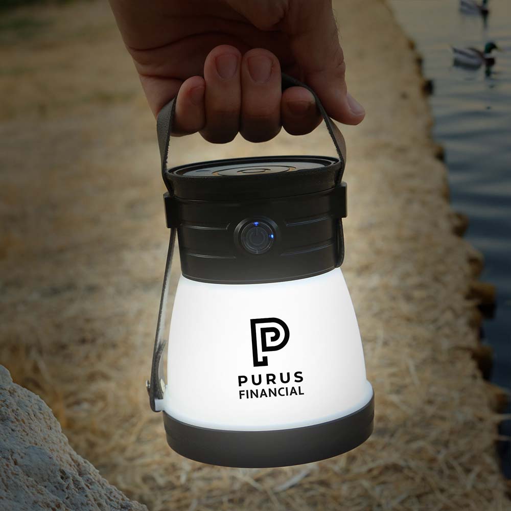 A hand holds a glowing LED lantern with a black handle and top; the white body features a customized logo in black