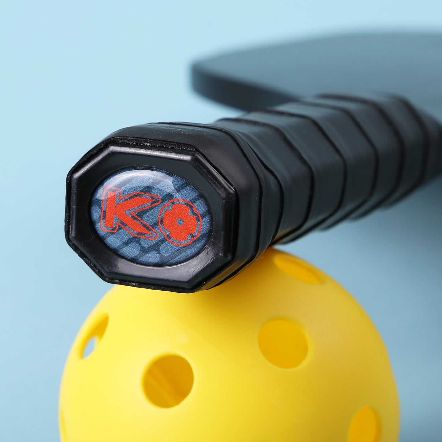 Close-up of a pickleball paddle handle with a textured black grip and a logo on the butt cap