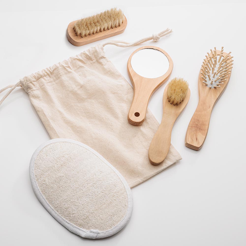 Natural spa kit with beige drawstring pouch, wooden-handled mirror, brushes, comb, and loofah pad — no logo imprint visible.