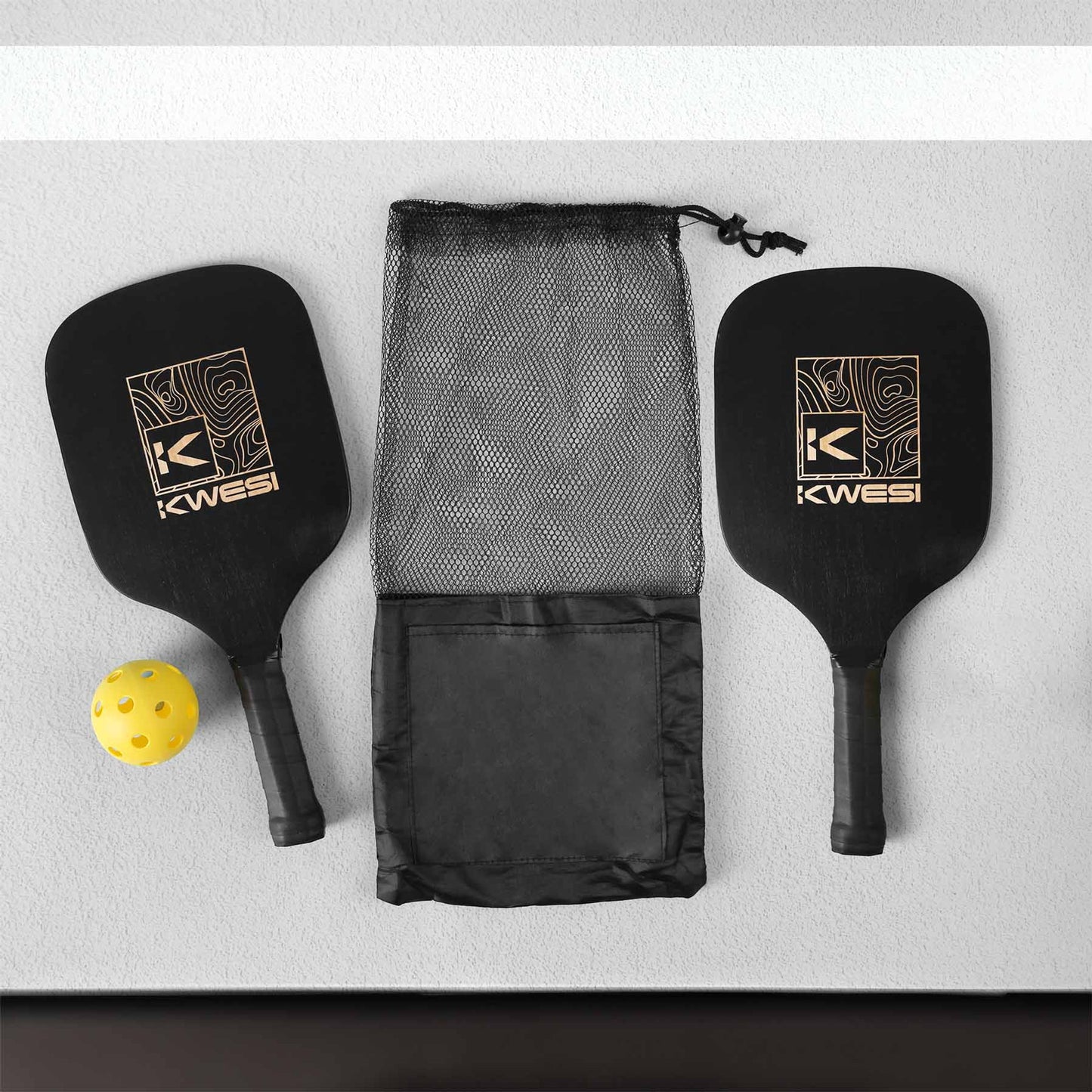 Pickleball set featuring two black paddles with logos, a yellow perforated ball, and a black mesh drawstring carrying bag with a front pocket