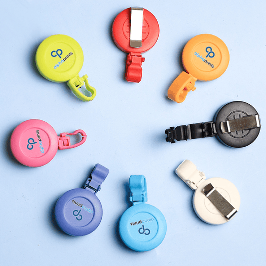 Assorted Custom Logo Printed Badge Reels in vibrant colors including red, orange, yellow, green, pink, purple, blue, black, and white, featuring clip attachments and the AlphaPrints logo, arranged in a circular pattern on a light blue background.