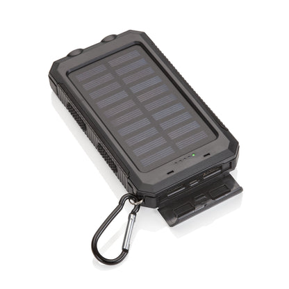 Angled view of a black solar power bank with a large solar panel, visible indicator lights, open protective port cover showing USB ports, and a metal carabiner attached to the corner.