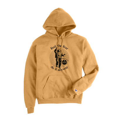 Golden brown Champion hoodie featuring a front graphic of a person standing with a bicycle inside a circular line pattern, accompanied by the motivational text “Feel the fear and do it anyway.” The Champion logo is embroidered on the left sleeve cuff.