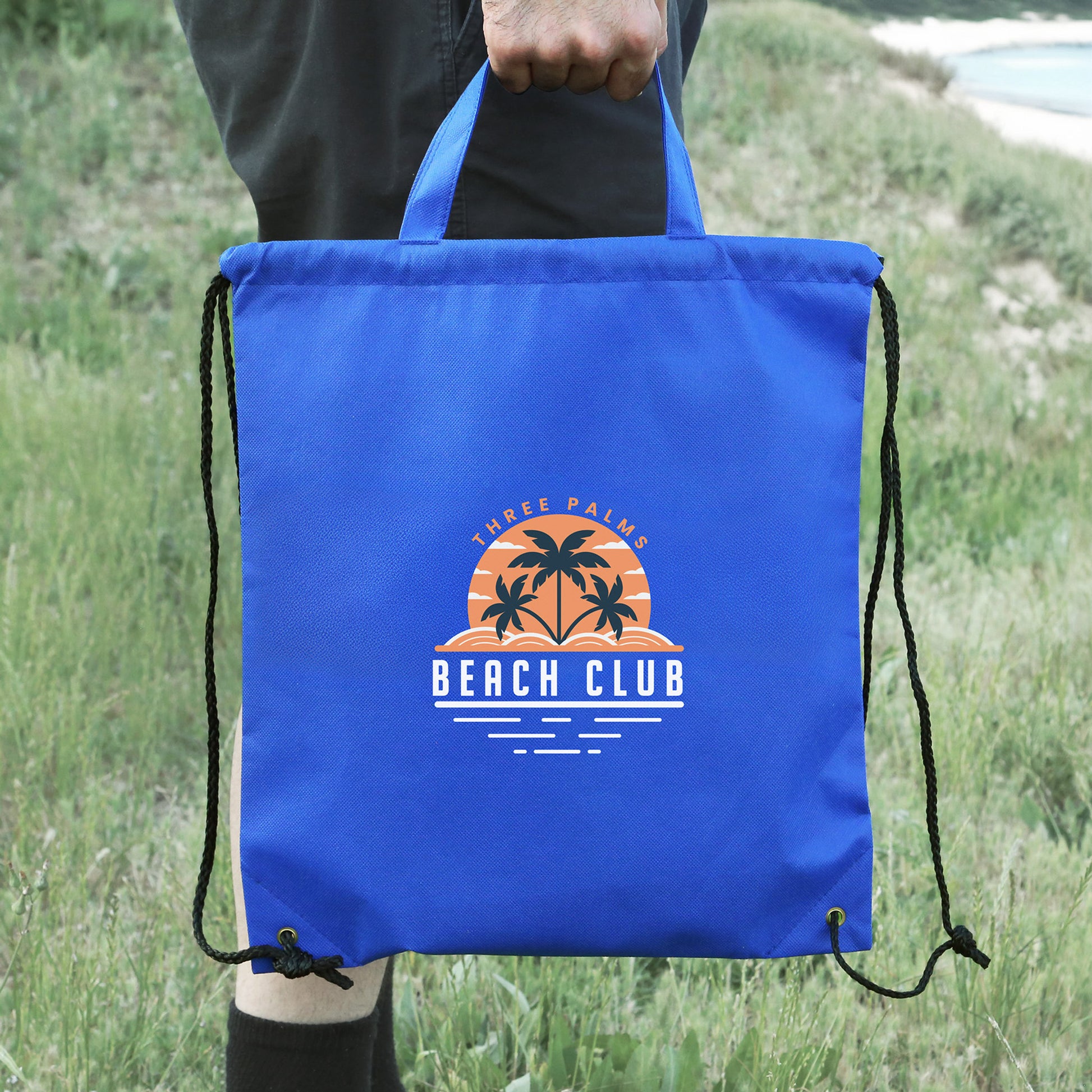 Blue Sportsman drawstring backpack with top handle, held outdoors by a person near grassy terrain, printed with designed logo